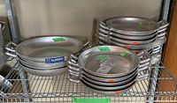 Stack of Italian aluminum saucepans with double handles, labeled with pricing and stacked on a wire shelf