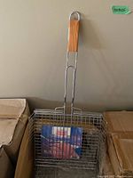 Photo of a single large BBQ grill basket with a wooden handle placed inside a box.