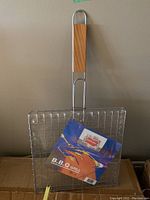 Two large BBQ grill baskets with wooden handles shown leaning against a wall with packaging label visible on front basket.