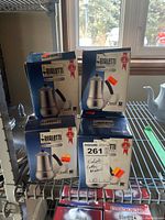 Twelve boxed Bialetti Elegance stainless steel coffee makers stacked on metal shelving, blue and white boxes with orange price stickers.