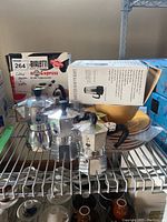 Four Bialetti coffee makers displayed on a metal shelf with packaging boxes behind, including cappuccino and espresso makers made in Italy.