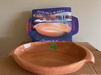 Front view of a terracotta ceramic baking dish with side handles and a green tag showing dimensions 13x8 inches, set against a cardboard box background.