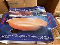 Top view of open box showing a terracotta-colored ceramic oval baking dish inside packaging with label and kitchen background imagery.