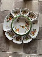 Top view of one set showing one large center platter surrounded by eight smaller plates with pomegranate and floral decoration on white ceramic