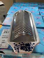 Top view of stainless steel cheese grater mounted on white glazed pottery dish with blue floral decoration on handle area.