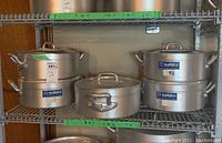Shelf holding five metal commercial cooking pots with lids, all marked Metalco and size 8 1/2 except one smaller pot in the center.
