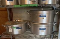 Three large aluminum cooking pots displayed on a metal shelving unit. Two pots stacked on the right side and one separate pot on the left, all with lids. Each pot has two side handles and a Metalco brand sticker.