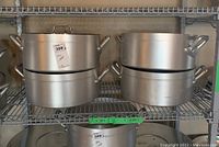 Four large aluminum cooking pots on shelf, three missing lids, with handles on each side.