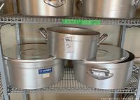 Three aluminum cooking pots on a shelf, two larger rectangular pots and one smaller round pot stacked on top, showing brand label and handles