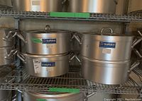 Four stacked heavy gauge aluminum pots with matching lids on a metal shelving unit, showing brand Metalco with LT14 model label.