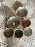 Six vintage Italian copper kitchen items including three bowls with brass handles, one colander, and two pans, arranged on a tiled floor.
