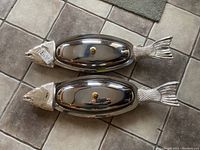 Overhead view of two fish-shaped platters with stainless steel lids showing fish head and tail handles.