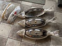 Three fish-shaped stainless steel platters arranged side by side, two with covers and one open, showing lids and overall shape.