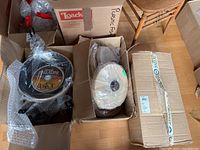 Boxes of new 12-inch Teflon coated frying pans with labels and packaging visible, showing three separate cardboard boxes containing pans wrapped in bubble wrap and plastic.