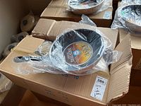 Six frying pans with lids in two open cardboard boxes, pans wrapped in plastic.