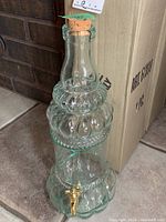 Two clear glass beverage dispensers, each approximately 15 inches tall, with cork stoppers and gold spigots. Dispensers have a textured segmented design and greenish tint.