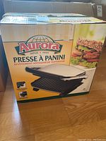 Boxed Aurora Panini Press grill showing front panel with product image and sandwich picture.
