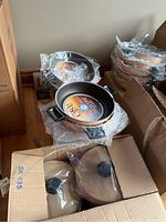 Photo showing multiple Teflon coated pans stacked, some wrapped in plastic, lids inside a box and stacked on the side.