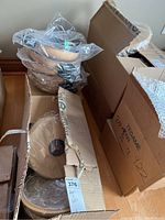 Six Ballarini 10x2 pans stacked and wrapped in clear plastic inside an open box, along with a box of lids.