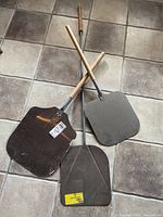 Three stainless steel pizza peels with wooden and metal handles, laid on tiled floor.