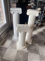 Three plastic Ionic style columns with fluted shafts and volute capitals, shown indoors on tile floor near glass doors and a fireplace.