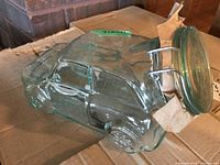 Clear pressed glass jar shaped like a vintage car with detailed molding and hinged clip-top lid with rubber seal, lying on its side on a cardboard box.