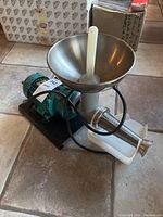 Electric countertop tomato press showing stainless steel funnel, plastic pusher, motor, and attached cords on tiled floor.