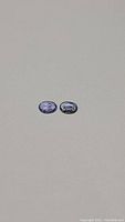 Pair of small oval cut tanzanite stones on a plain surface, showing blue-purple color and oval shape.
