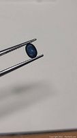Photo showing natural oval cut blue sapphire held by tweezers against a plain background.