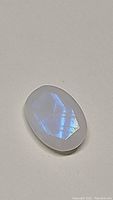 Oval cut rainbow moonstone gemstone showing translucent white color with blue flash on a neutral background.