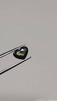 Close-up image of a tri-coloured heart-shaped sapphire gemstone held by tweezers, showcasing green, yellow and blue hues.