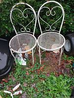 Pair of vintage white metal garden chairs with heart-shaped backrest and circular mesh seat, shown outdoors on grass with some rust and paint chipping.
