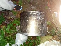 Side exterior photo of the aluminum pot lying on grass, showing wear, dirt, and surface scratches.