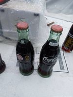 Two green tint glass Coca-Cola bottles with red caps, 1996 Olympics logo visible on one bottle, both are dusty and unopened.