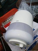 Photo showing a white and translucent plastic towel dispenser with a knob at the front. Brand label 'Tork' visible at the top center of the dispenser.