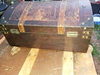 Side view of a small wooden chest with curved top, metal corner reinforcements, leather straps, and metal hardware.