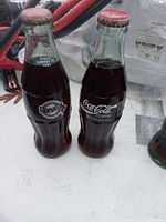 Two unopened vintage Coca-Cola glass bottles with Super Bowl logos visible on labels, showing rounded classic Coca-Cola bottle shape and red caps.