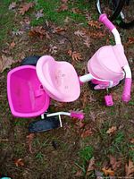 Photo of a pink and white children's tricycle on grass. Shows molded pink seat, back storage bin, front canopy, and pink pedals. The item appears dirty and in need of cleaning.