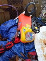 Side and angled views of orange and yellow plastic ride-on toy tow truck with black steering wheel and rear hook, lying on tarp near fence with some leaves on and around it.