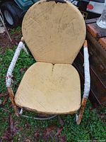 Image shows a single vintage chair with round backrest and square seat. Upholstery is yellow and heavily soiled, with visible scratches. Metal armrests are wrapped partially with white material that is deteriorating. The chair is positioned outdoors on grass and leaves.