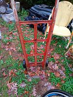 Photo of a single vintage metal hand truck standing on grass with dry leaves, showing red paint and black wheels.