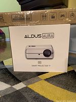 Front view showing white Aldus Aura projector box and long cardboard projection screen package