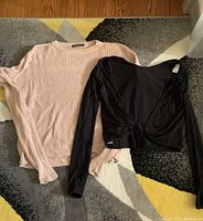 Two tops laid flat on the floor: light pink ribbed sweater from Brandy Melville and black long-sleeve knot-front top from Wilfred
