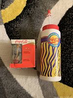 Photo of Coca-Cola mug next to collectible bottle in original box on carpet surface.