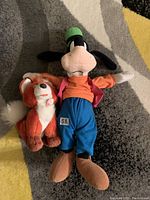 Photo showing the front view of Goofy and Tod plush toys laid on a carpet, Goofy wearing colorful clothes and Tod with a white tipped tail.