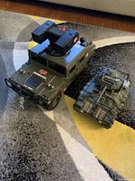 Two military toy vehicles on floor carpet: GI Joe branded Humvee with missile launcher and unbranded toy tank vehicle.
