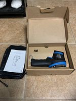 Infrared thermometer tool in box with instruction manual and carrying case laid nearby on tiled floor.