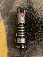 Close-up photo of the Yoda lightsaber hilt showing the metal clip, red button, and wear marks.