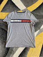 Full view of the grey Tommy Hilfiger T-shirt laid flat on patterned carpet showing the horizontal logo print across the chest.