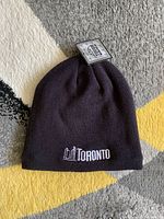 Black knit beanie hat laid flat on a multi-colored carpet, showing the Toronto skyline embroidery and attached tag